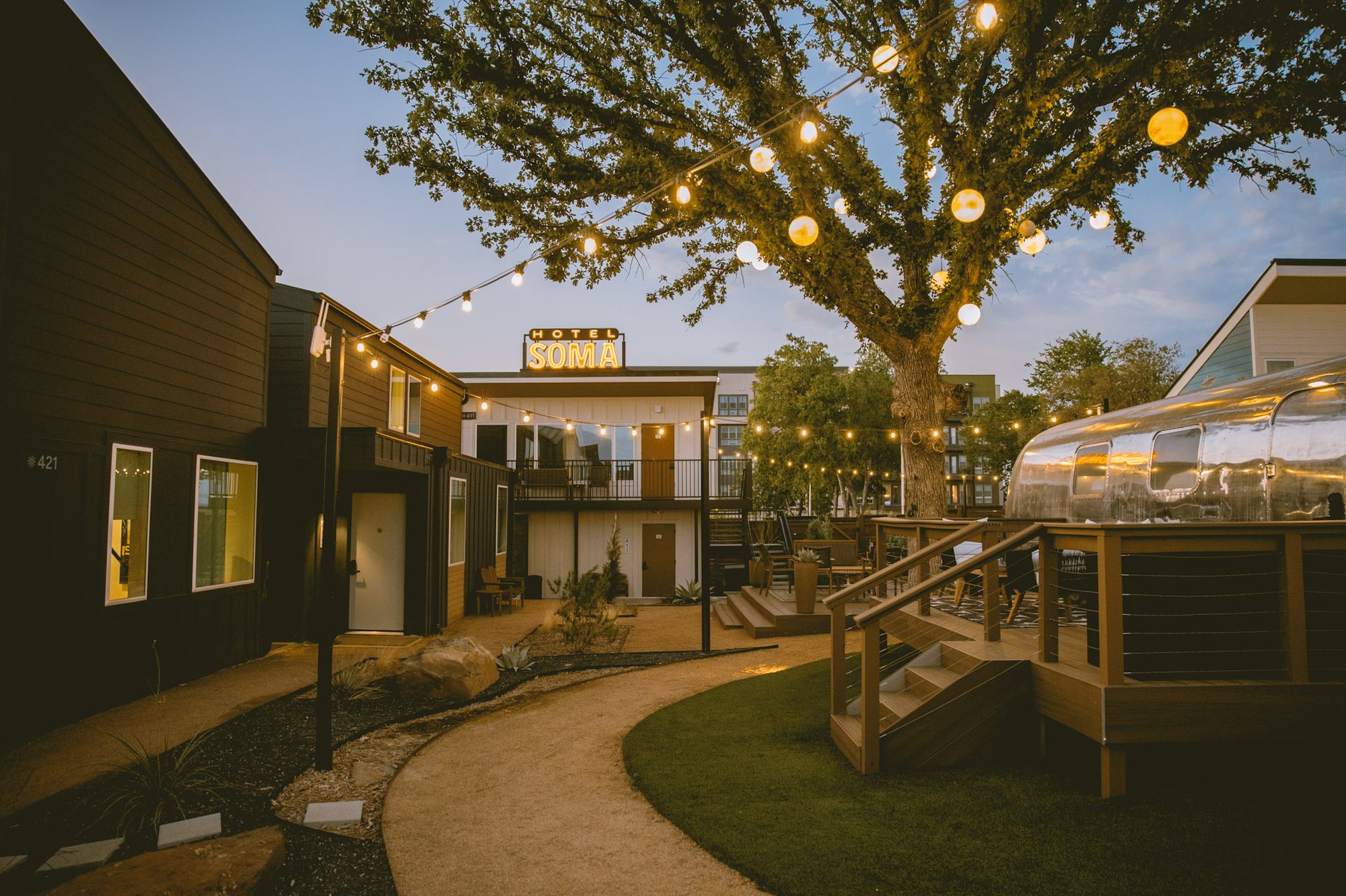 Golden-hour light over the Hotel SOMA courtyard, string lights and Airstream suites beneath the oak in Fort Worth